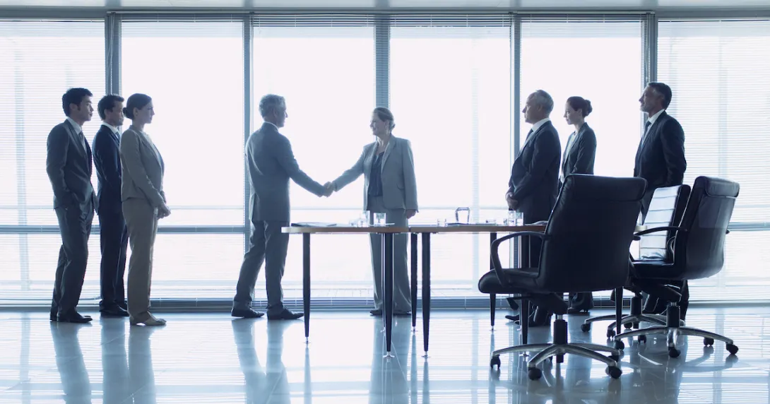 People standing in front of a large window with two of them shaking hands and six others standing and watching