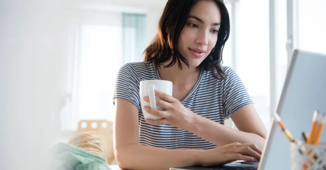   Person in a gray shirt sitting down while holding a coffee mug and looking at a computer